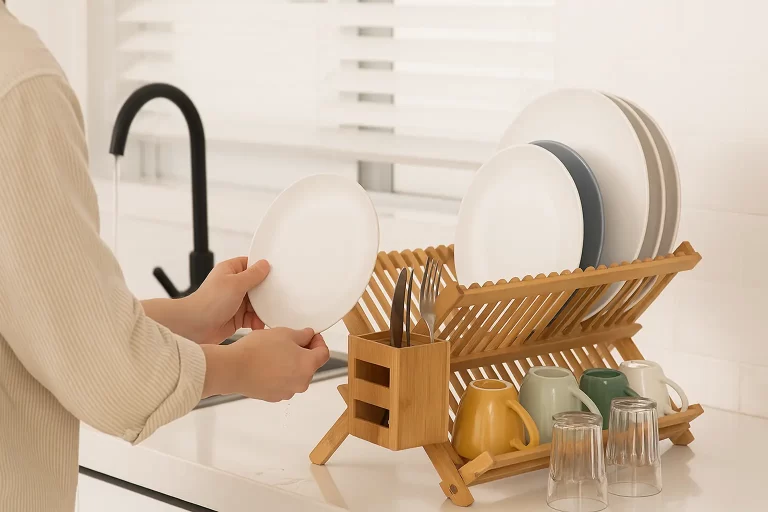 A person placing a clean plate on a Bamboo Dish Drying Rack, with coffee cups, glasses, and utensils neatly drying in a bright modern kitchen